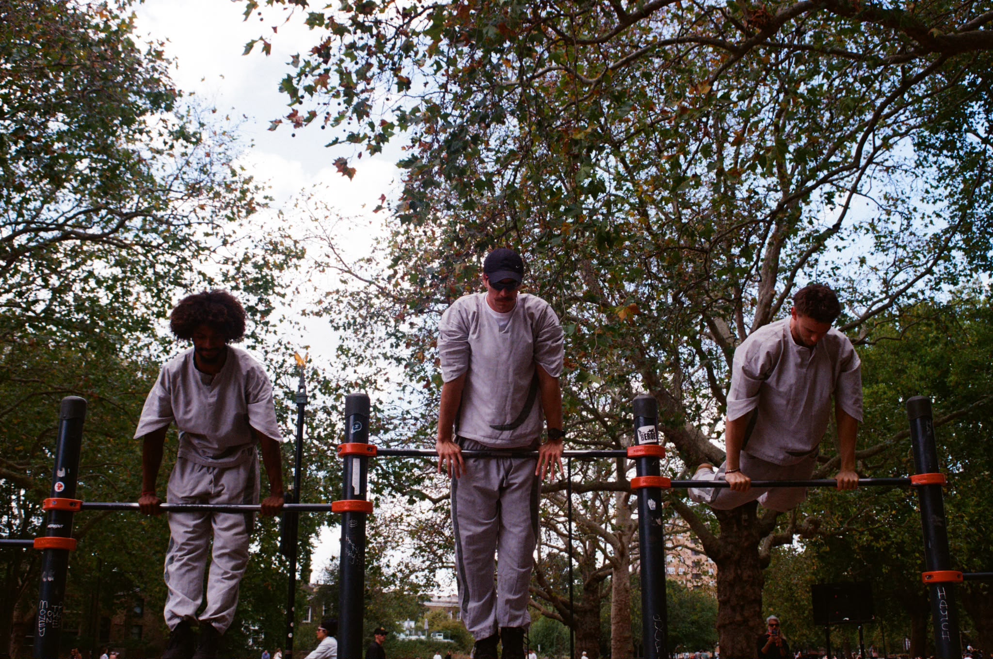 Three people holding a support position on top of parallel bars in a park, wearing light grey hemp sets.