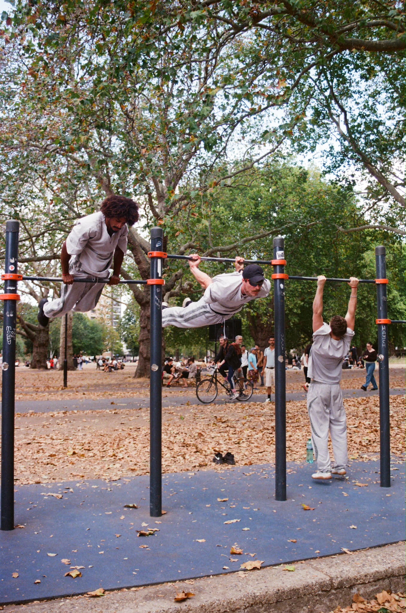 Three people training calisthenics on outdoor bars — one in a muscle-up, one in a front lever, one in a pull-up.