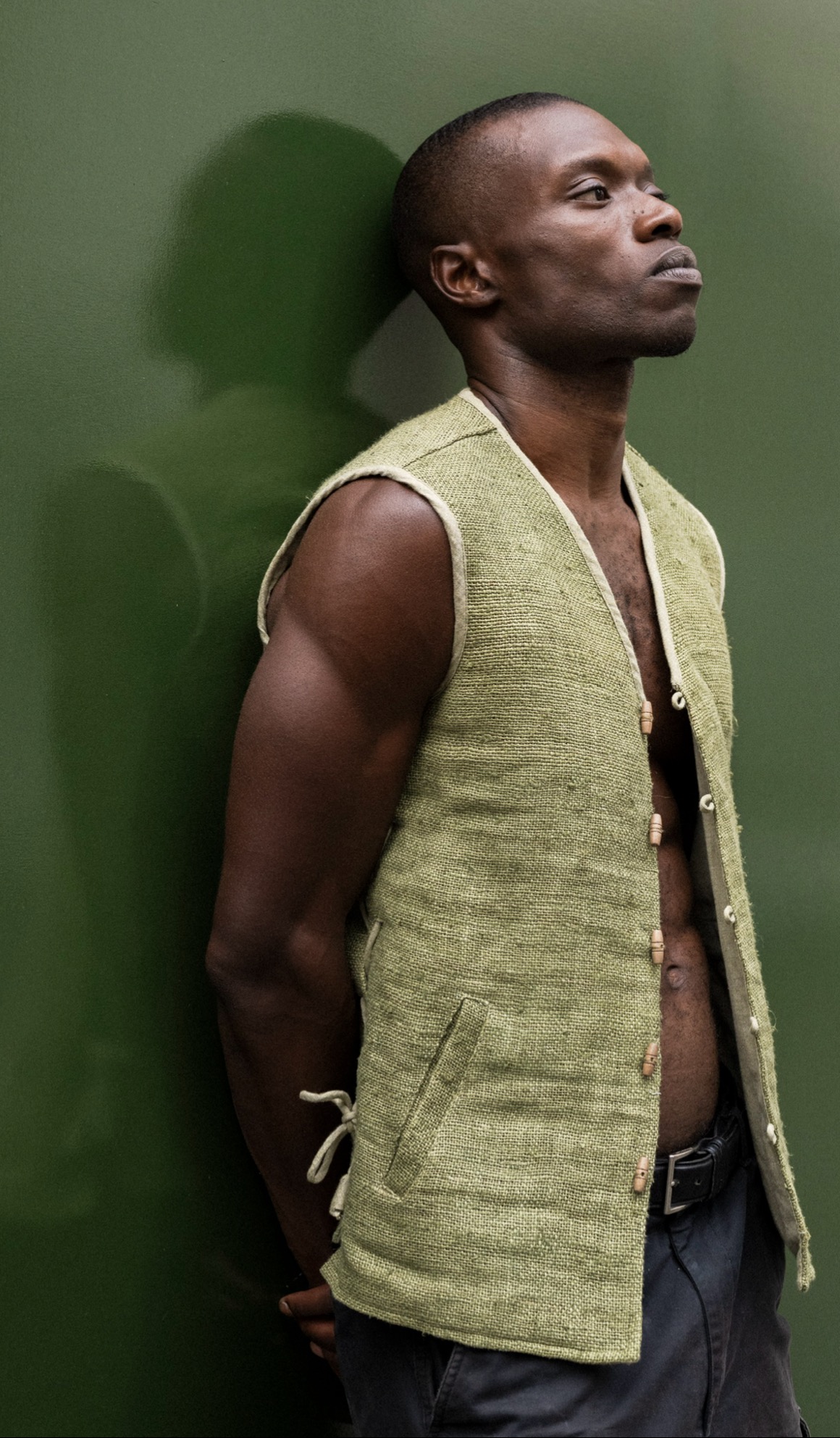 Man in profile wearing a sage woven hemp vest with wooden toggles, standing against a deep green wall